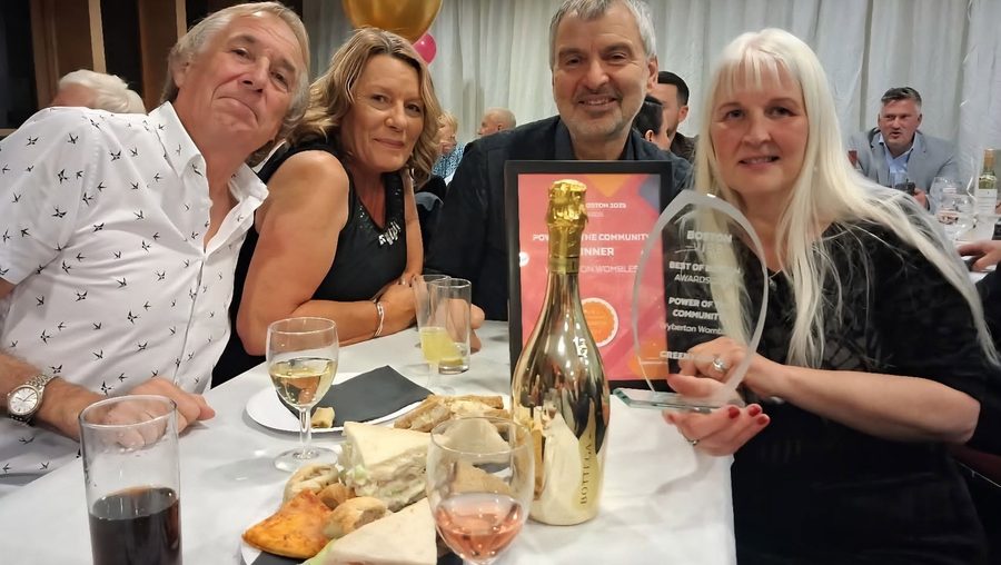 Four smiling people at a dinner table with a framed certificate and a glass trophy that read Power of the Community, Wyberton Wombles