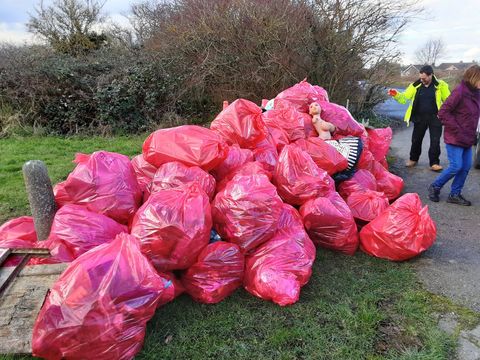 A huge heap of full red sacks on a grass verge at a roadside, with two volunteers standing beside it