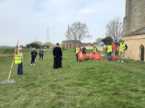 Volunteers in hi-vis picking litter on the green beside a stone church, with a vicar in a black cassock standing among them and red sacks on the grass