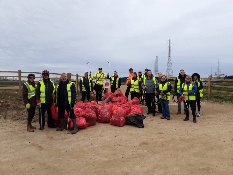 A large group of volunteers in hi-vis vests with litter pickers, standing behind a big pile of full red sacks on a dirt track, with a wooden fence and electricity pylons behind