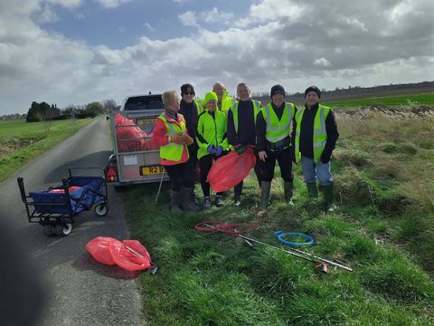 Seven volunteers in hi-vis vests standing on a grass verge beside a country lane, next to a trailer and a pull-along cart, with red sacks and litter pickers on the grass