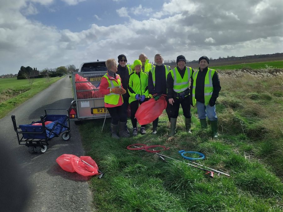 Seven volunteers in hi-vis vests standing on a grass verge beside a country lane, next to a trailer and a pull-along cart, with red sacks and litter pickers on the grass