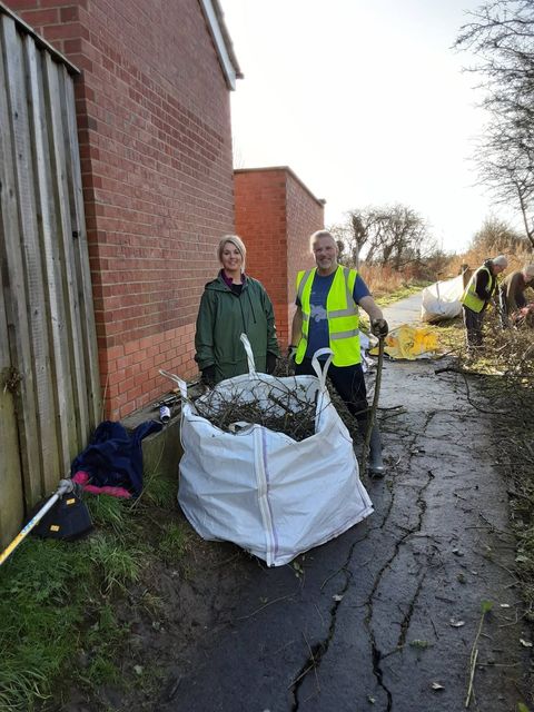 Two volunteers standing in an alleyway between brick walls beside a large white bulk bag full of rubbish, with a litter picker and red sacks nearby