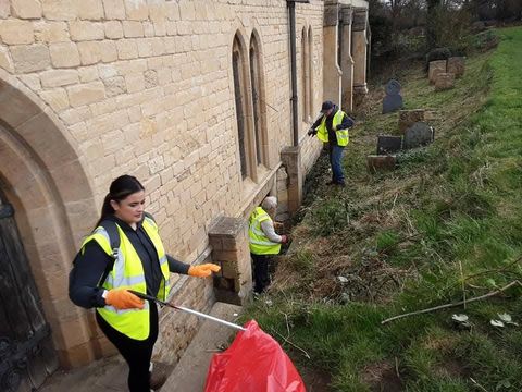 A smiling volunteer in a hi-vis vest and orange gloves holding a litter picker and red sack beside a stone church wall, with two more volunteers working among the gravestones behind